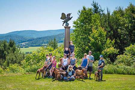 A group of people and dogs posing in front of a scuplture by Stephen Huneck of an angel dog on a sunny day with hills and trees in the background
