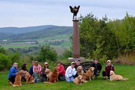 People posing together with their dogs at an outlook over rolling green hills with a pillar on the right side that has an angel dog statue on top of it.