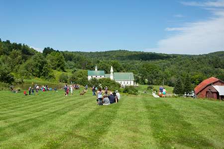 Dogs and their owners in a field and near a pond with a dog church and a bard in the background